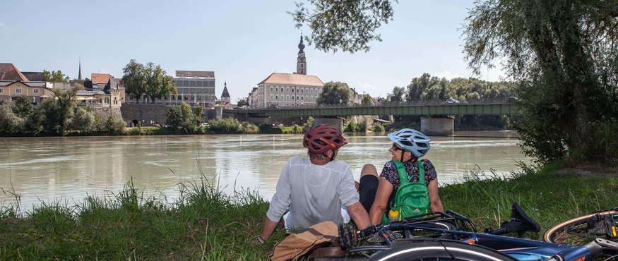 Two cyclists sitting by riverbank overlooking town and bridge