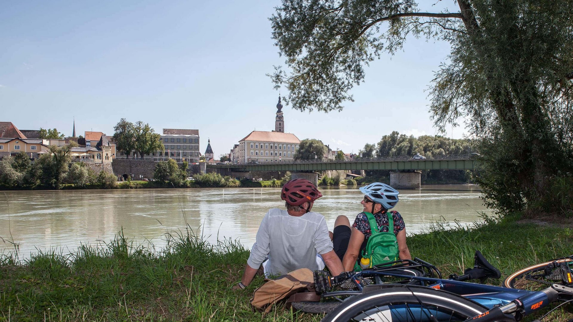 Two cyclists sitting by riverbank overlooking town and bridge