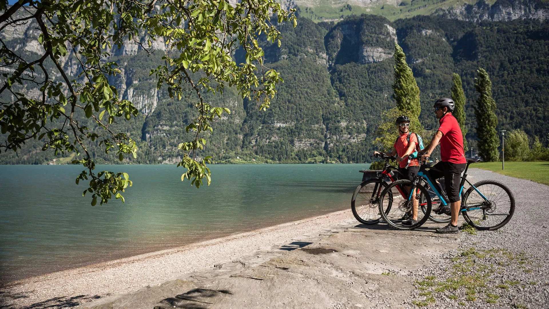 5 Punkte für Heidiland-Bucket List © Velontour Zwei Radfahrer am Seeufer mit Bergen im Hintergrund bei sonnigem Wetter
