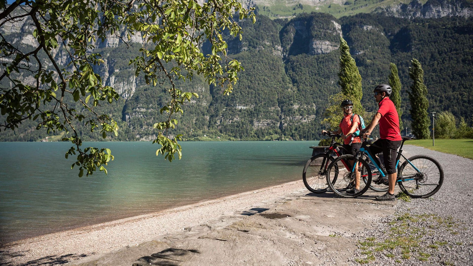 Zwei Radfahrer am Seeufer mit Bergen im Hintergrund bei sonnigem Wetter
