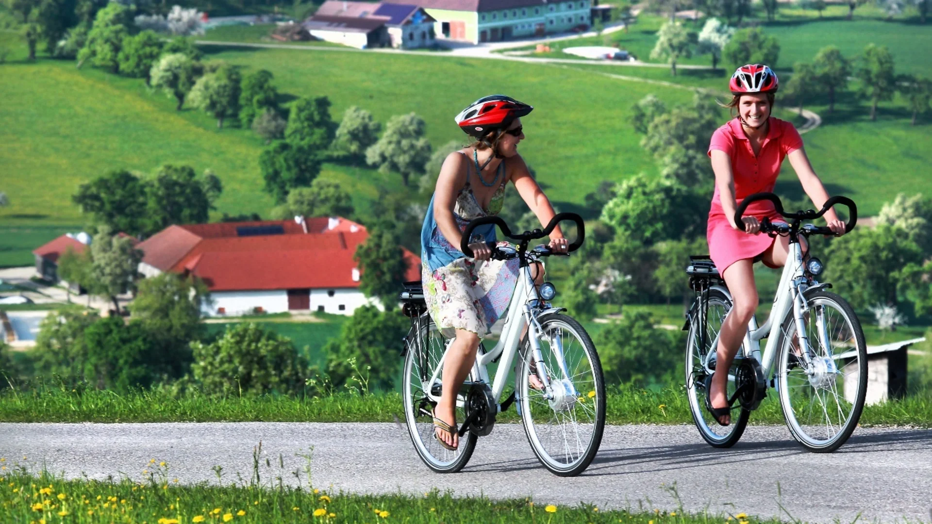 Tour-Detail Zwei Frauen mit Helmen fahren Fahrräder auf Landstraße mit grünen Feldern im Hintergrund