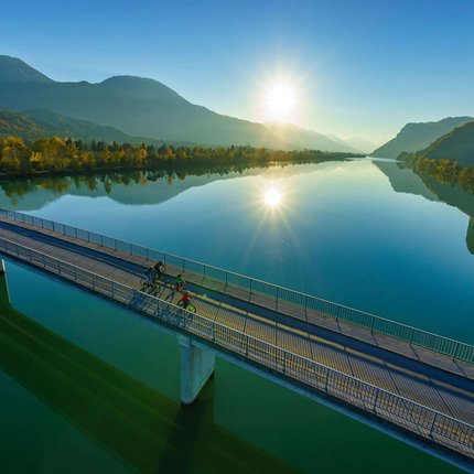 Cyclists on bridge over calm river at sunrise in mountainous landscape