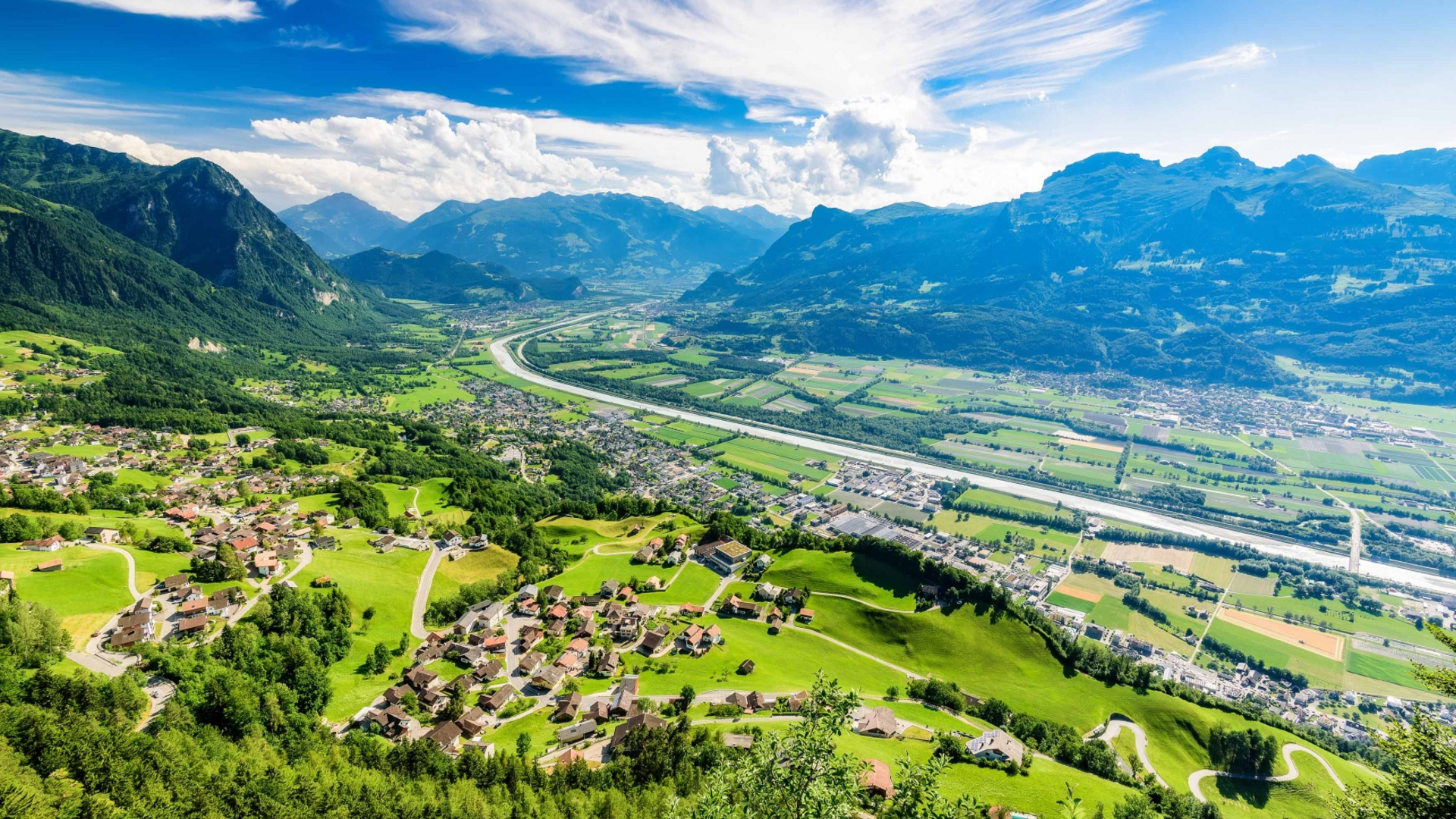 Aerial view of a valley with mountains, river, and villages under blue sky