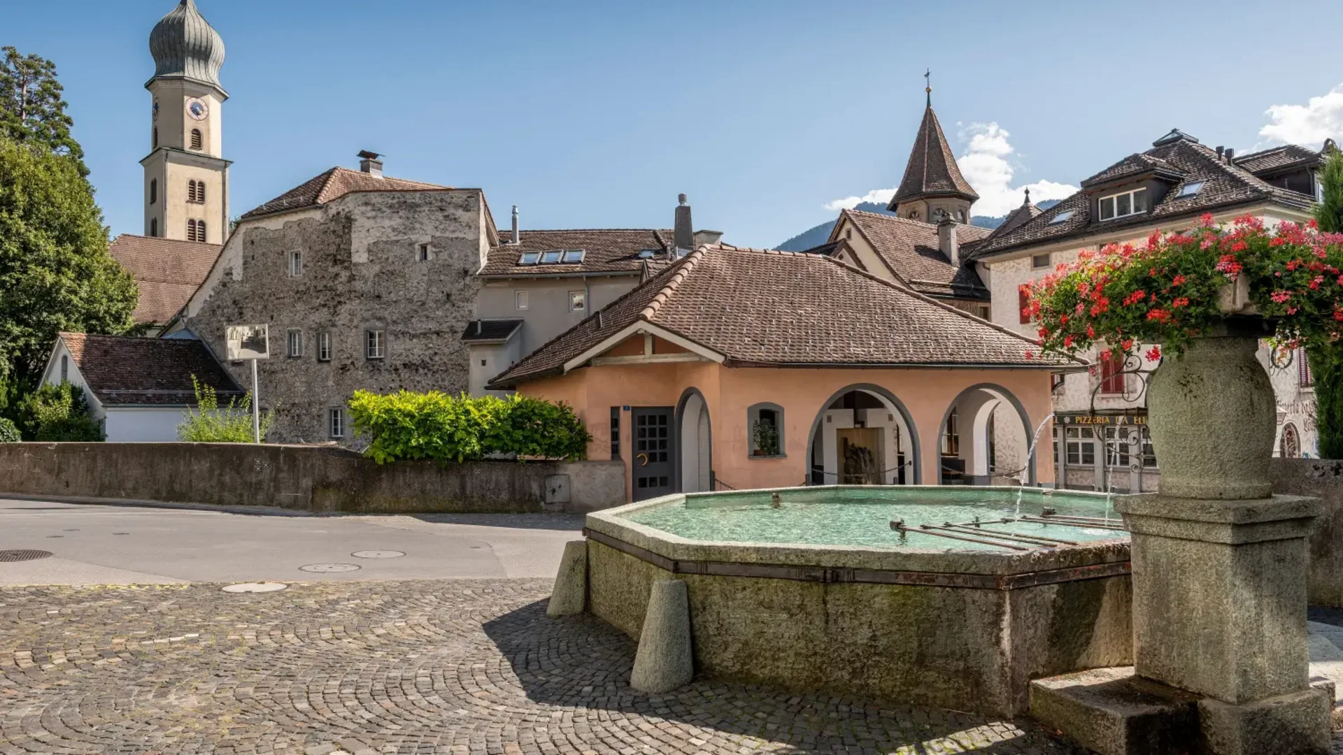 Historische Gebäude und ein Brunnen in einem sonnigen Dorf unter blauem Himmel
