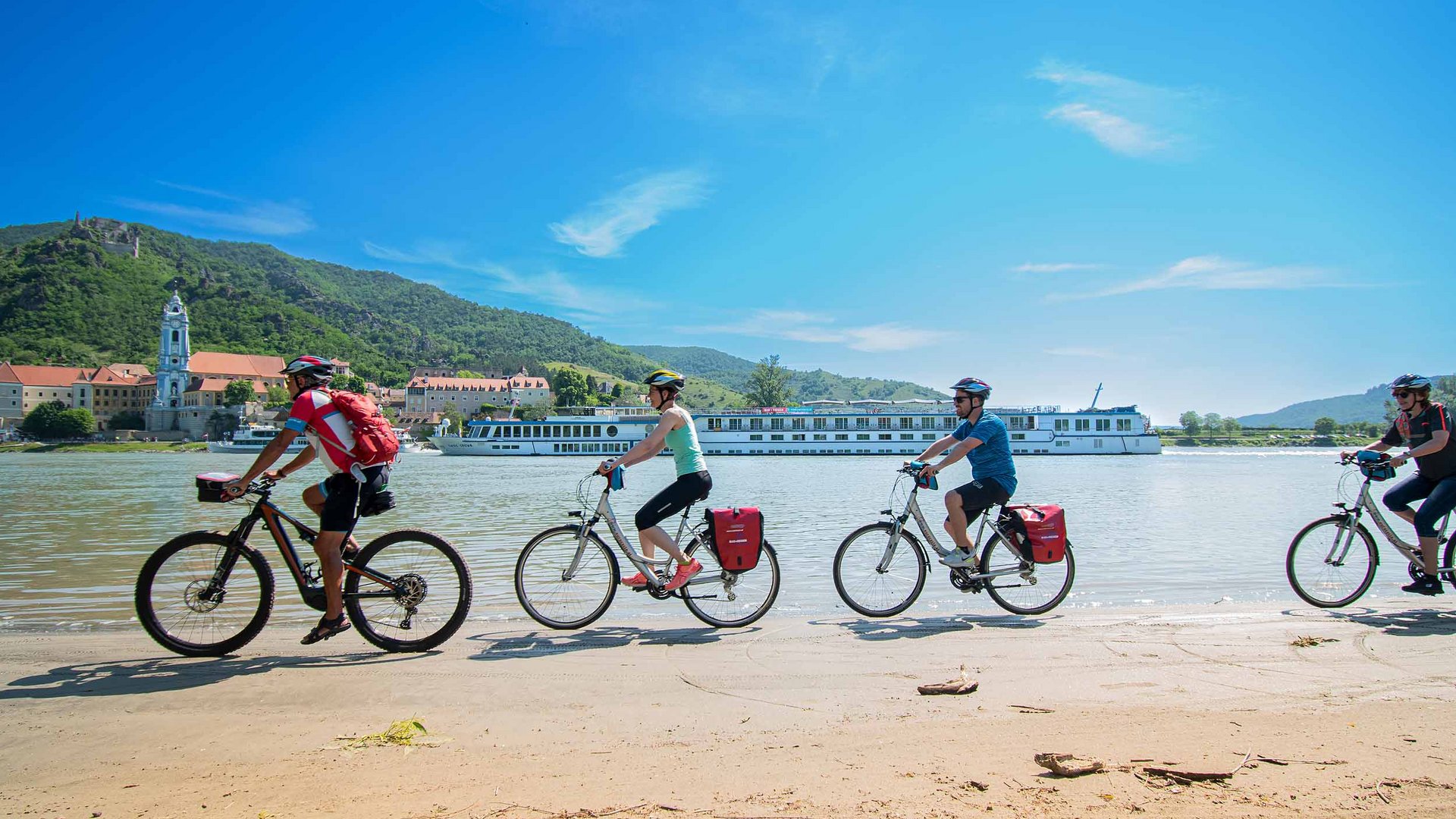 Four cyclists riding along a riverbank with a cruise boat in the background