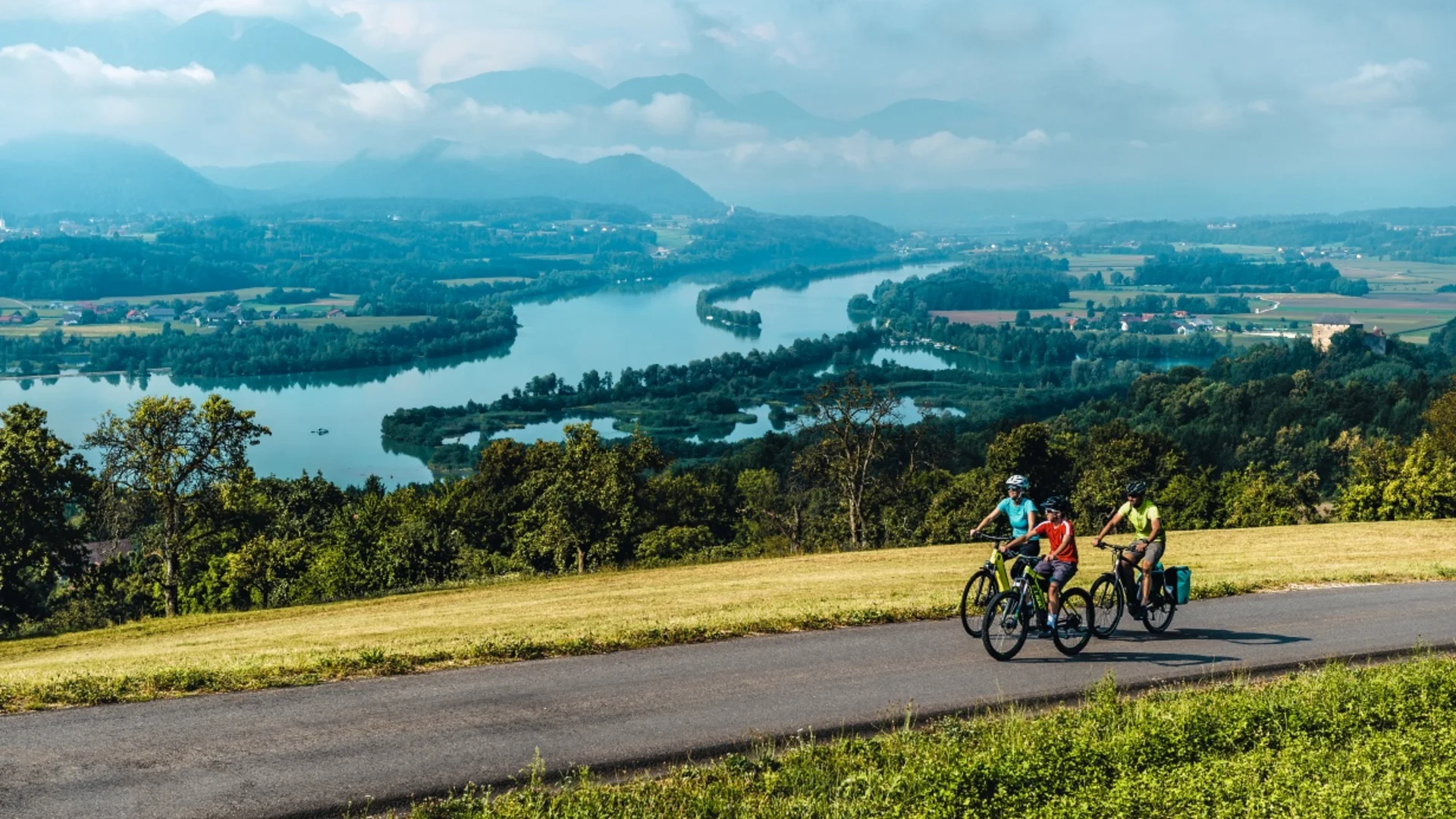 Drie fietsers rijden op een landweg met rivier en bergen op de achtergrond