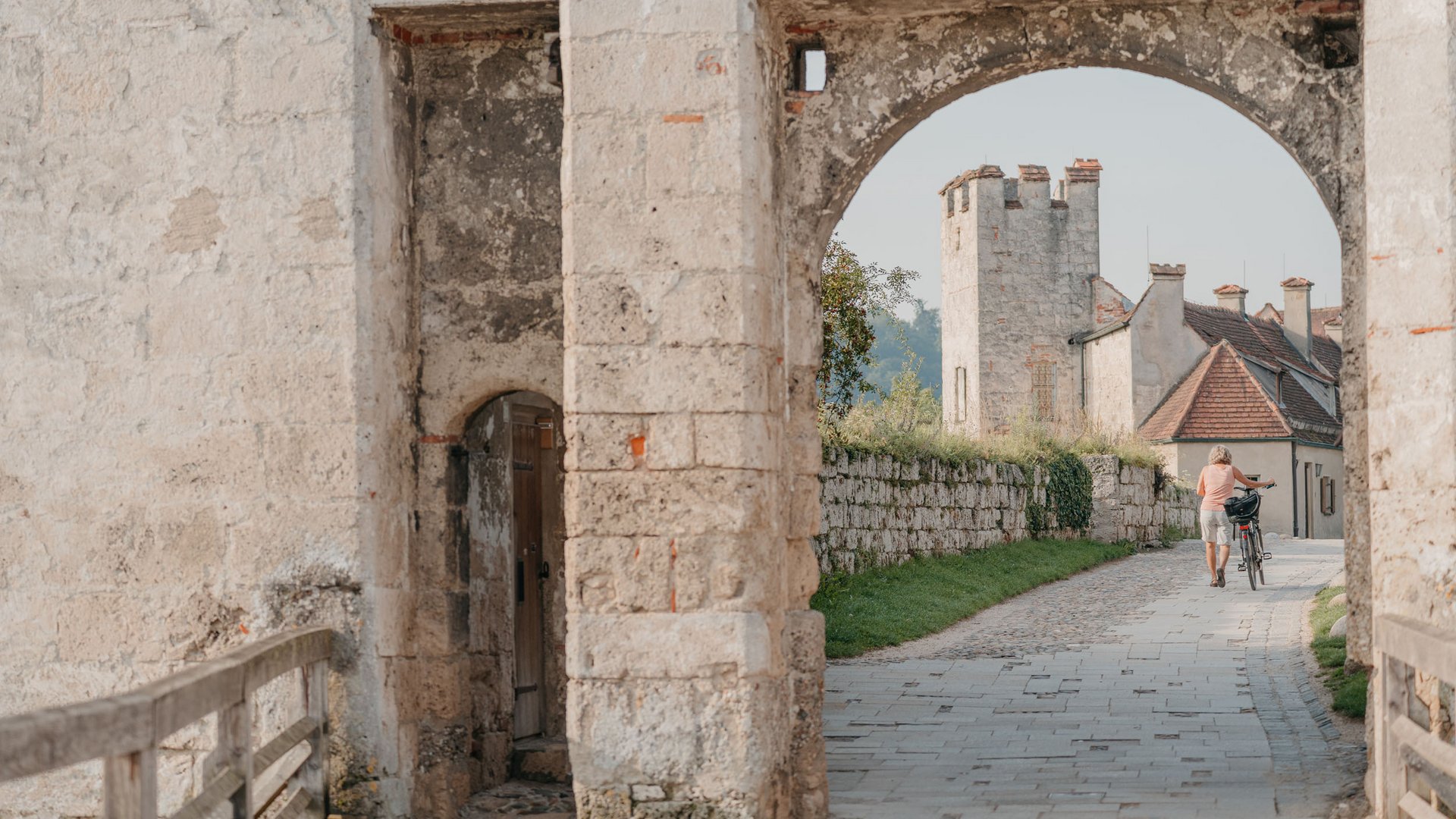 Person walking bike through old stone archway with castle in background