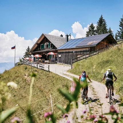 Twee fietsers op bergpad bij berghut met zonnepanelen op zonnige dag