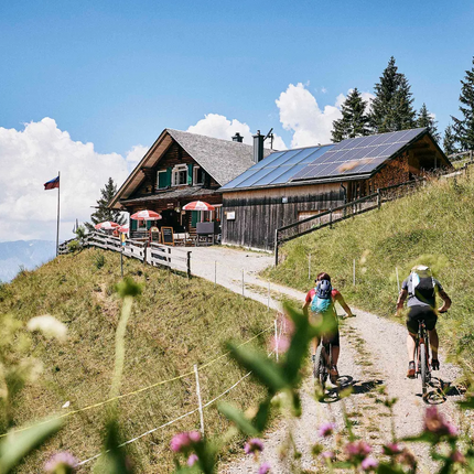 Two cyclists on mountain path near cabin with solar panels on sunny day