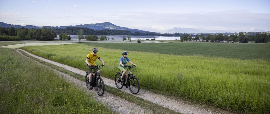 Two cyclists riding on a dirt path with green fields and a lake in the background