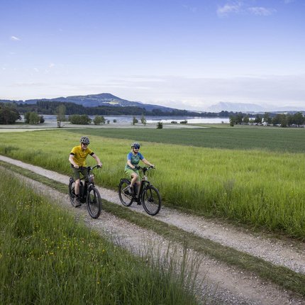 Two cyclists riding on a dirt path with green fields and a lake in the background