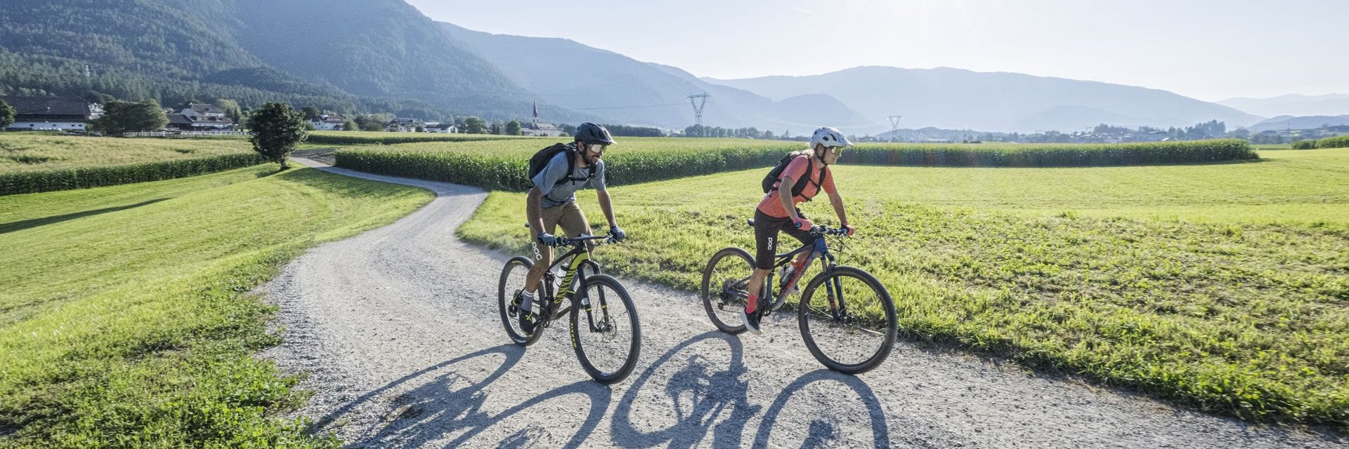 Two cyclists riding on a gravel path through sunny fields with mountains in the background