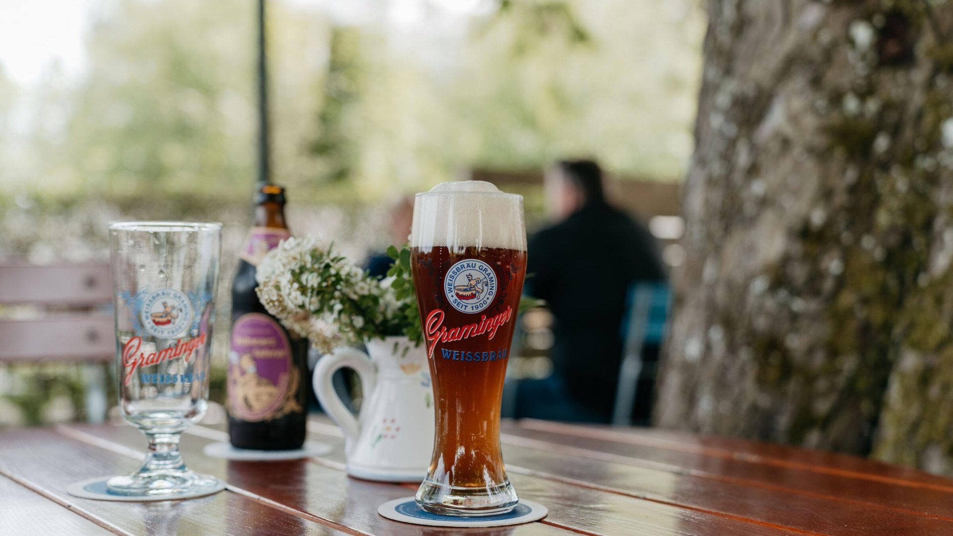 A glass of dark beer with foam on a wooden table next to flowers and a beer bottle