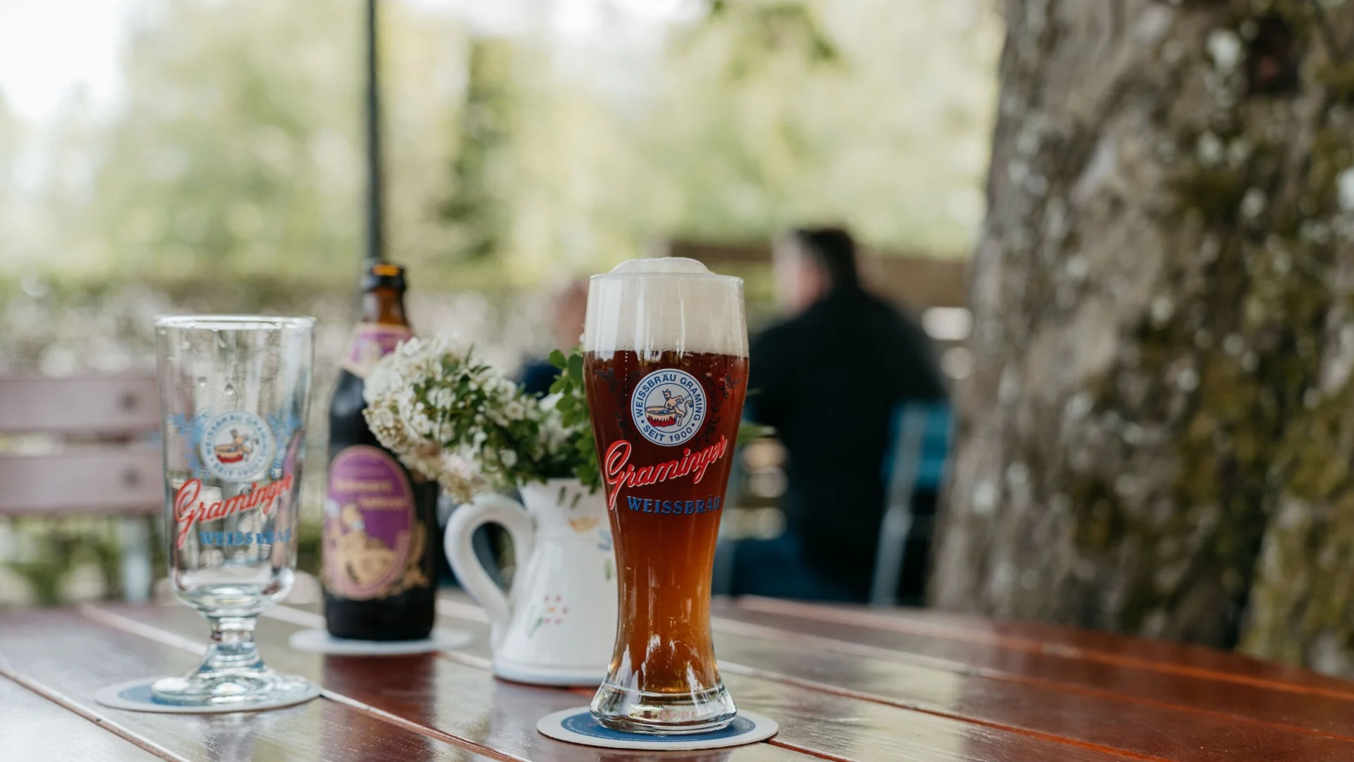 Ein Glas dunkles Bier mit Schaum auf einem Holztisch neben einer Blume und Bierflasche