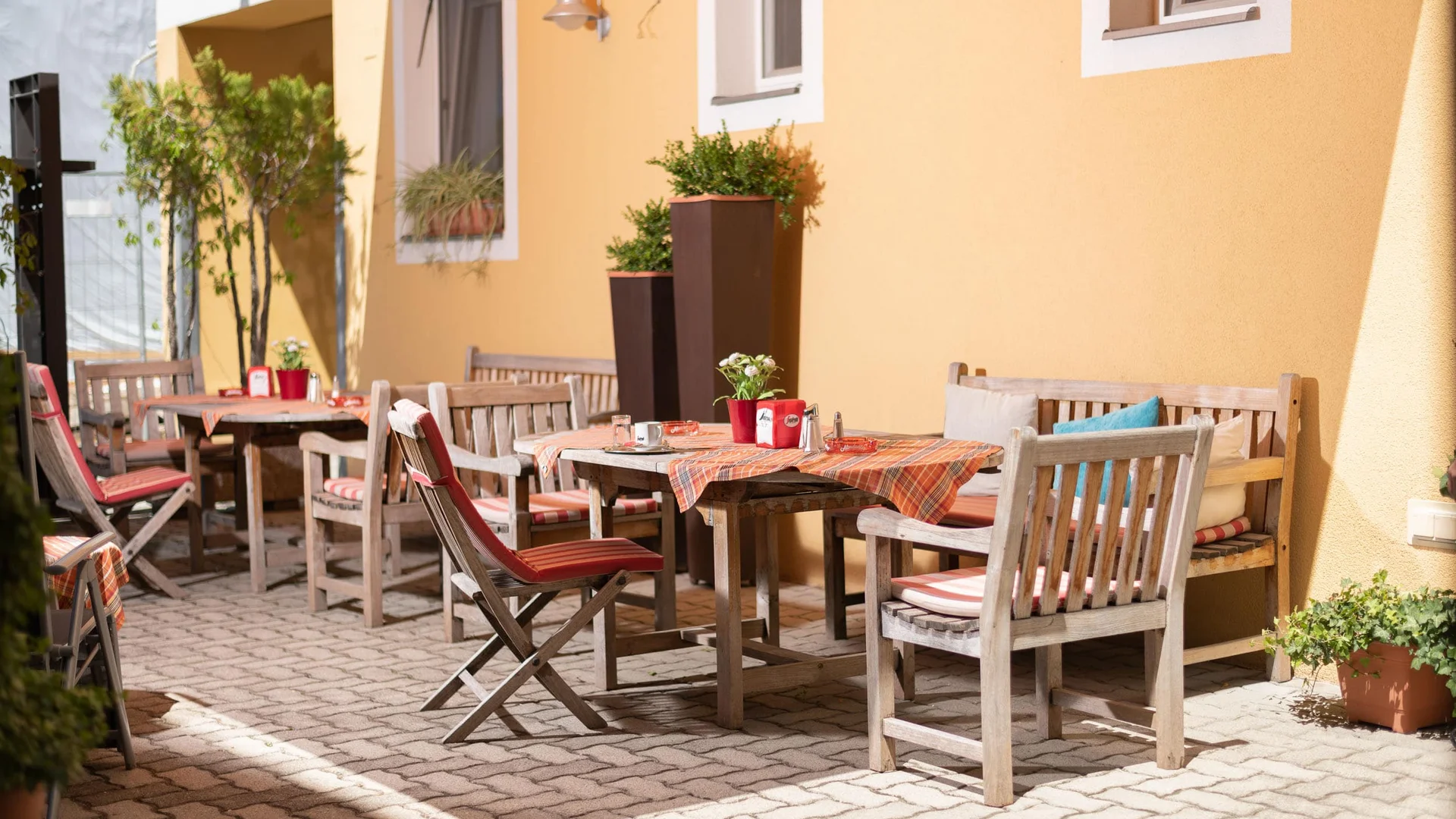 Zonnig buitenterras met houten tafels en stoelen bij gele muur