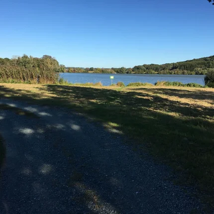 Gravelweg am See mit Bäumen und Hügeln unter klarem blauem Himmel