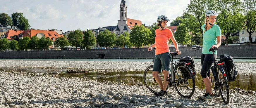 Zwei Frauen mit Fahrrädern stehen an einem Flussufer vor einer Kirche
