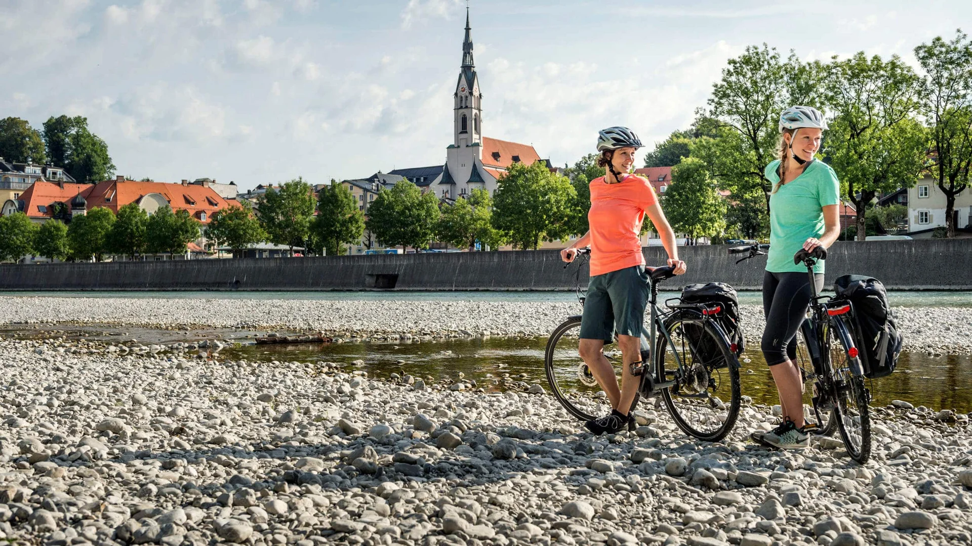 Zwei Frauen mit Fahrrädern stehen an einem Flussufer vor einer Kirche