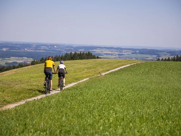 Zwei Radfahrer fahren auf einem Pfad durch grüne Hügel mit weitem Ausblick