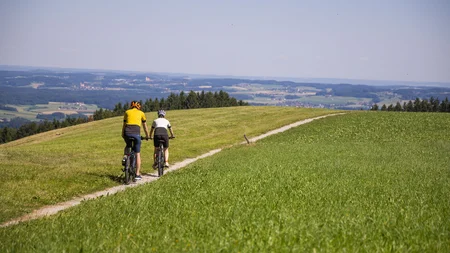 Zwei Radfahrer fahren auf einem Pfad durch grüne Hügel mit weitem Ausblick