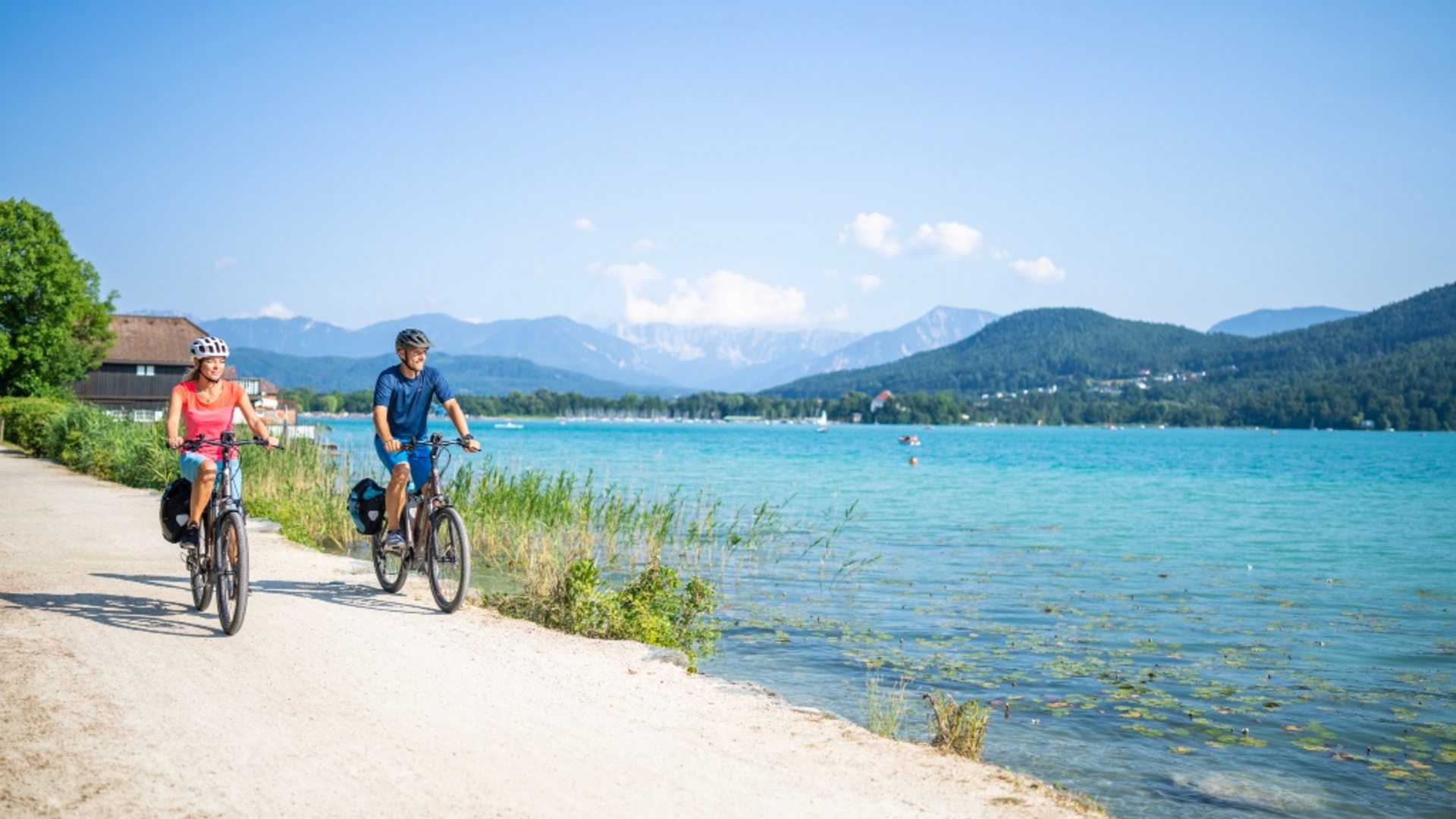 Two cyclists riding on a path by a lake with mountains in the background
