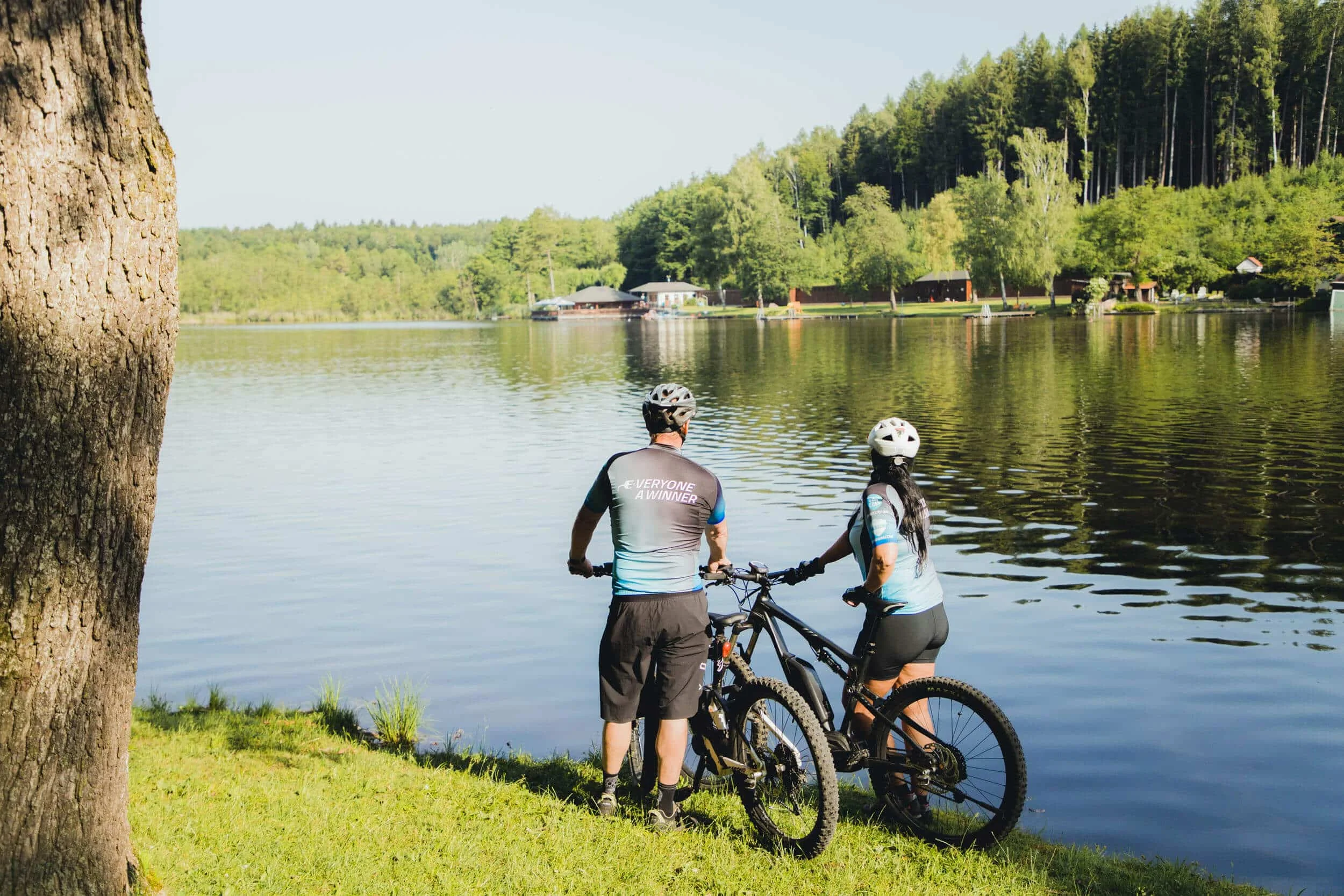 Zwei Radfahrer stehen am Seeufer und schauen auf den Wald und das Wasser