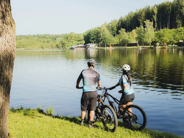 7 Quellen der Entschleunigung © E-Bike World Federation Zwei Radfahrer stehen am Seeufer und schauen auf den Wald und das Wasser
