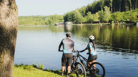 7 Quellen der Entschleunigung © E-Bike World Federation Zwei Radfahrer stehen am Seeufer und schauen auf den Wald und das Wasser
