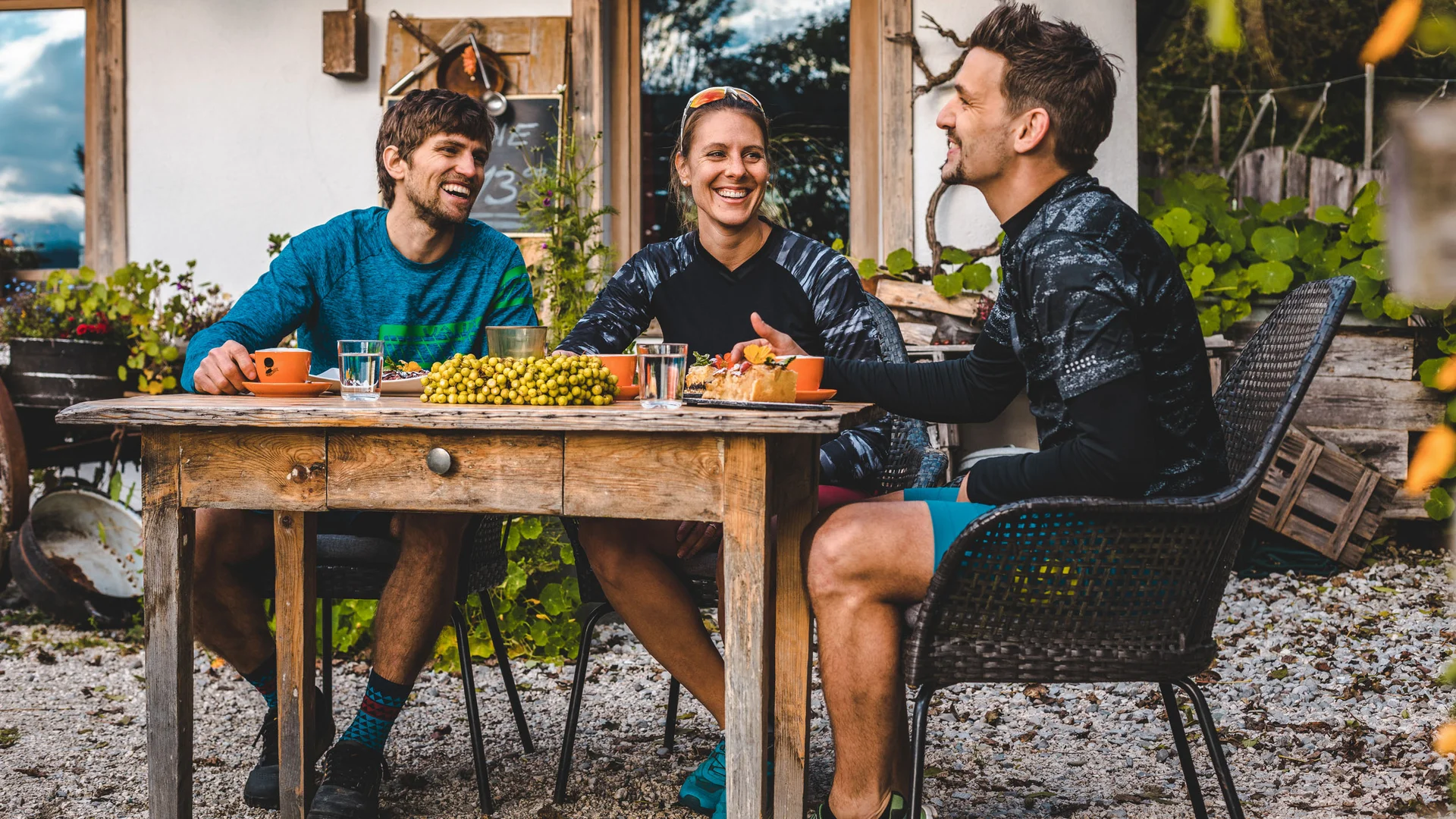 Drei Freunde sitzen draußen am Holztisch und lachen bei Kaffee und Kuchen