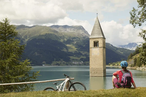 3 Highlights für den Radurlaub am Reschensee © IDM Südtirol - Matt Cherubino Fahrradfahrerin sitzt am See mit versunkenem Kirchturm und Bergen im Hintergrund