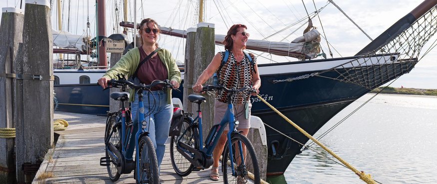 Two women with bicycles on a wooden pier next to a sailboat