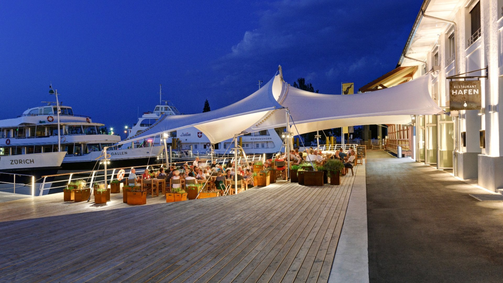Outdoor dining at Hafen restaurant by harbor with docked ships at dusk