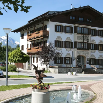 Traditionelles Gasthaus mit Brunnen und Statue vor Alpenkulisse