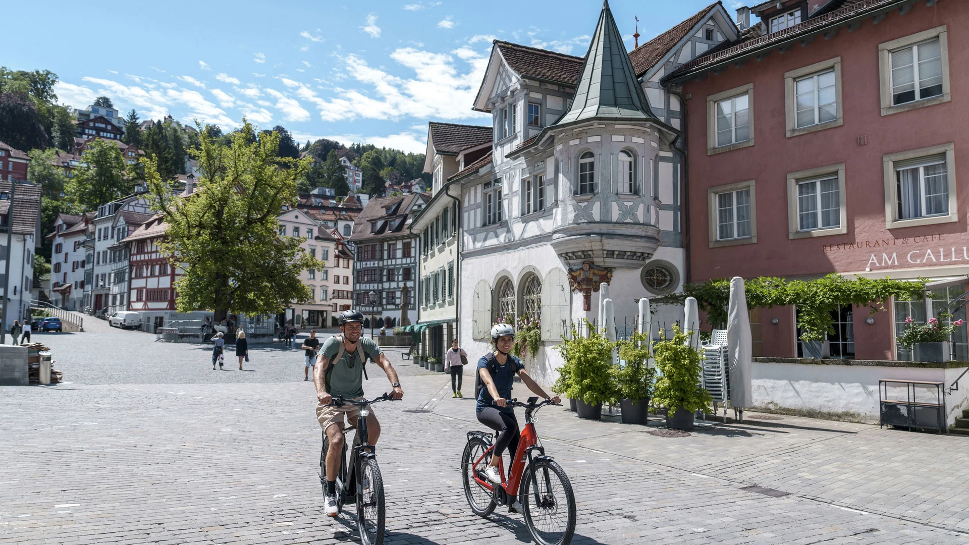 Twee mensen fietsen op een zonnige dag door een oud stadsplein