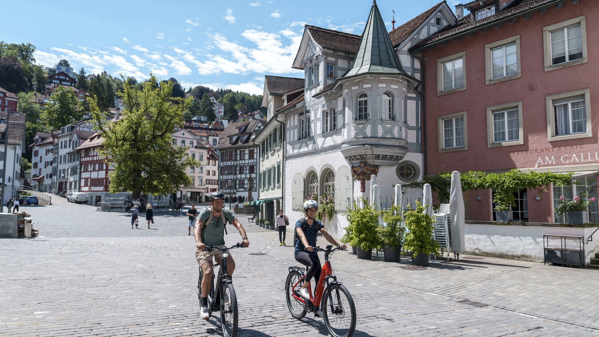 Two people riding bikes on a sunny day through an old town square