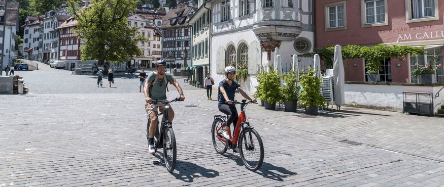 Two people riding bikes on a sunny day through an old town square