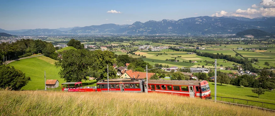 Rode trein rijdt door groen landschap met bergen onder een heldere lucht