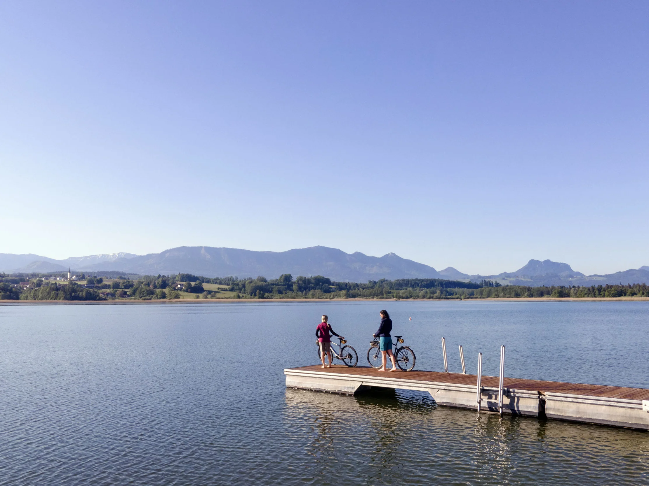 Deutschland © Oberbayern.de Zwei Personen mit Fahrrädern auf einem Steg an einem See mit Bergen im Hintergrund