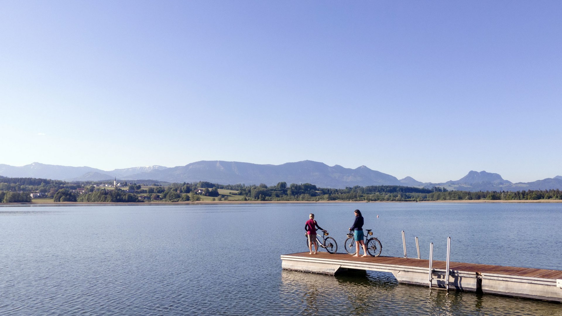 Two people with bicycles on a dock by a lake with mountains in the background