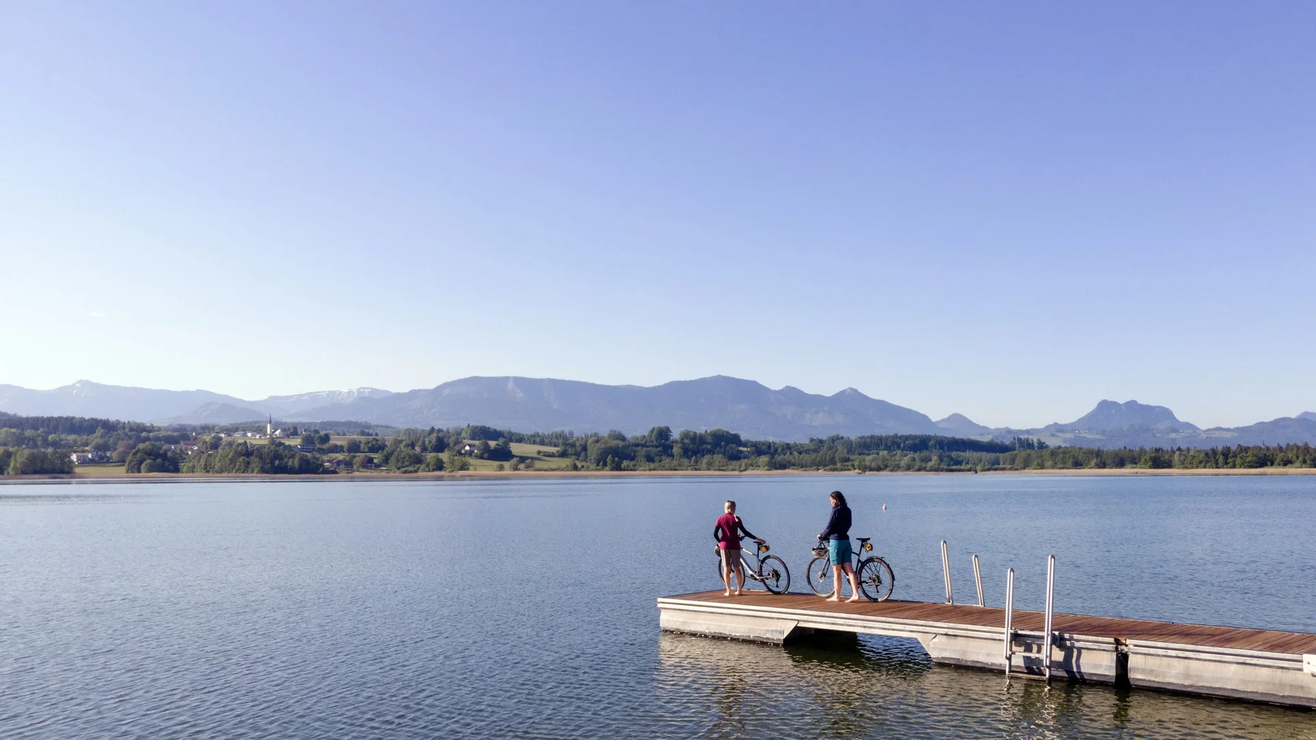 Twee personen met fietsen op een steiger bij een meer met bergen op de achtergrond