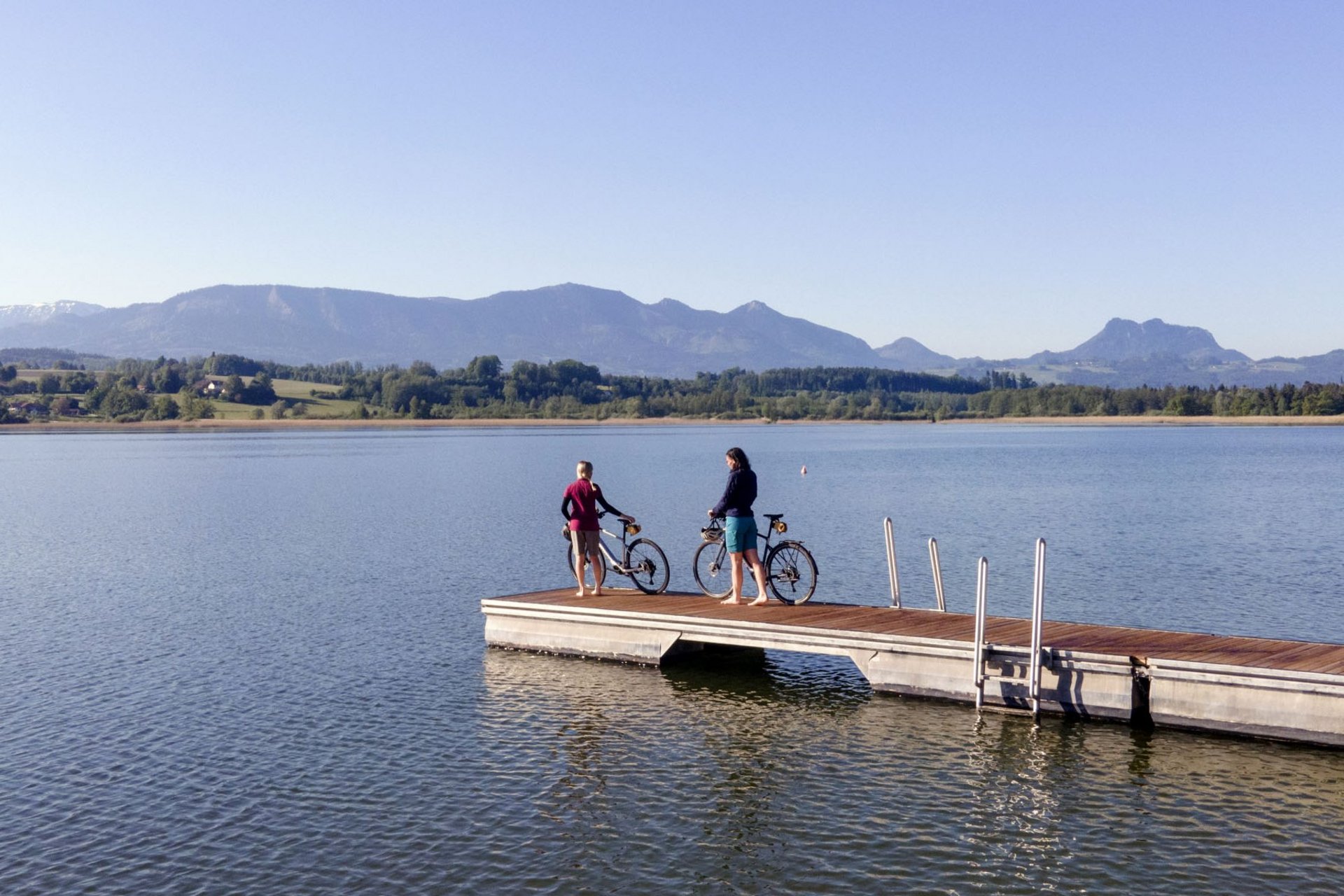 Two people with bicycles on a dock by a lake with mountains in the background