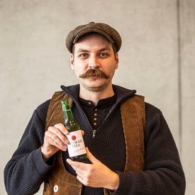 Mann mit Schnurrbart hält eine Flasche Pilsbier in Traditioneller Kleidung