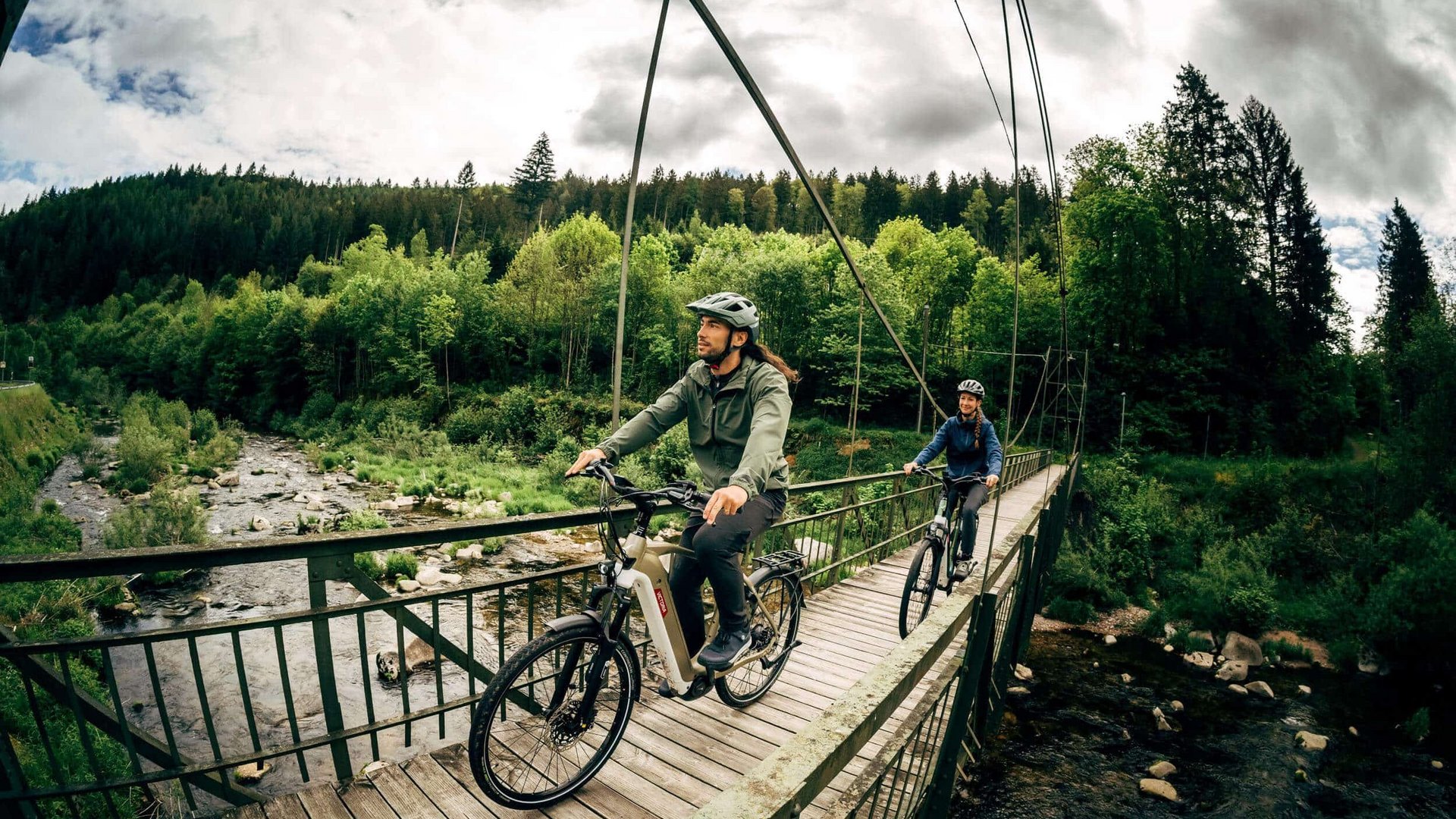 Victoria Two cyclists wearing helmets crossing a suspension bridge over a river in green nature