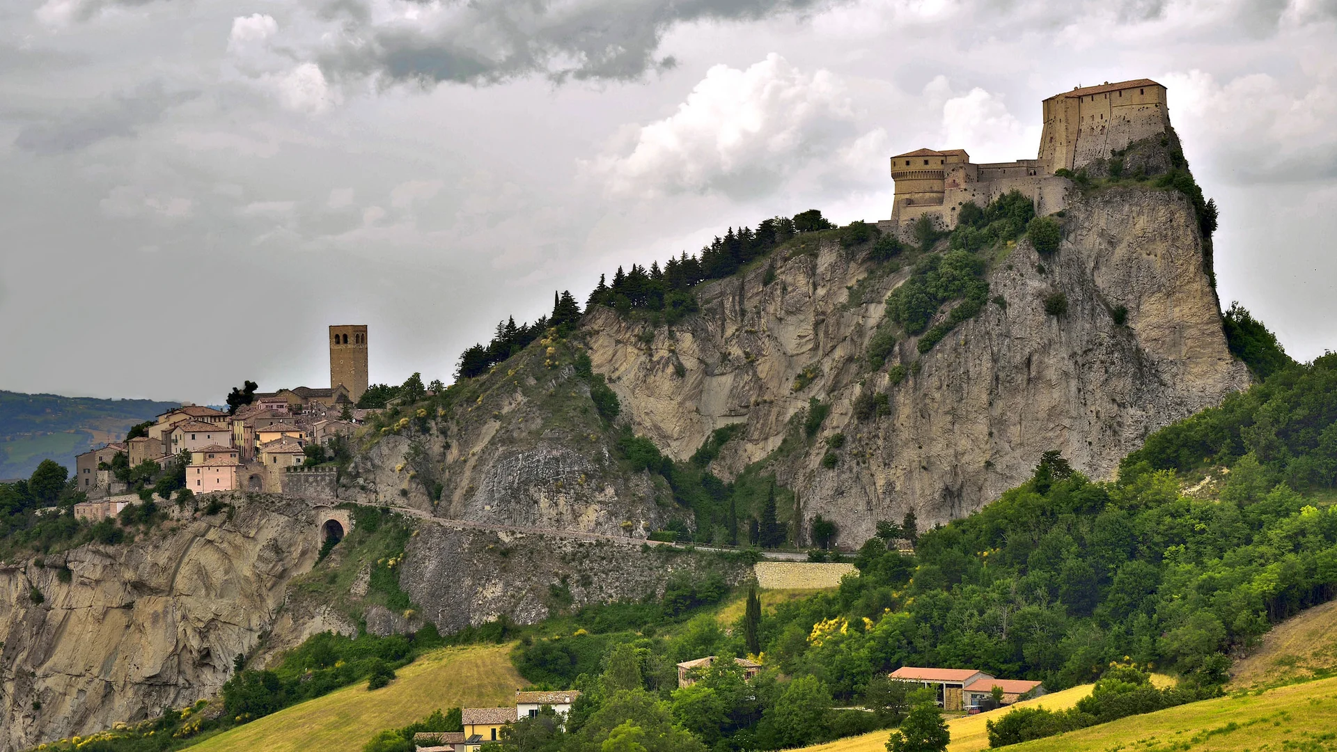 Festung und Dorf auf einem steilen Felsen mit grüner Landschaft und bewölktem Himmel