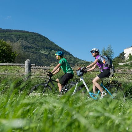 Two cyclists riding on a country path with a castle and mountains in the background