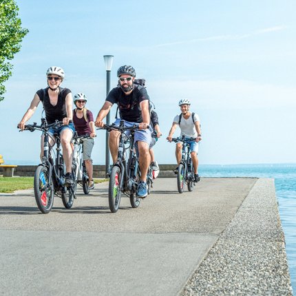 Group of cyclists wearing helmets riding along a waterfront path on sunny day