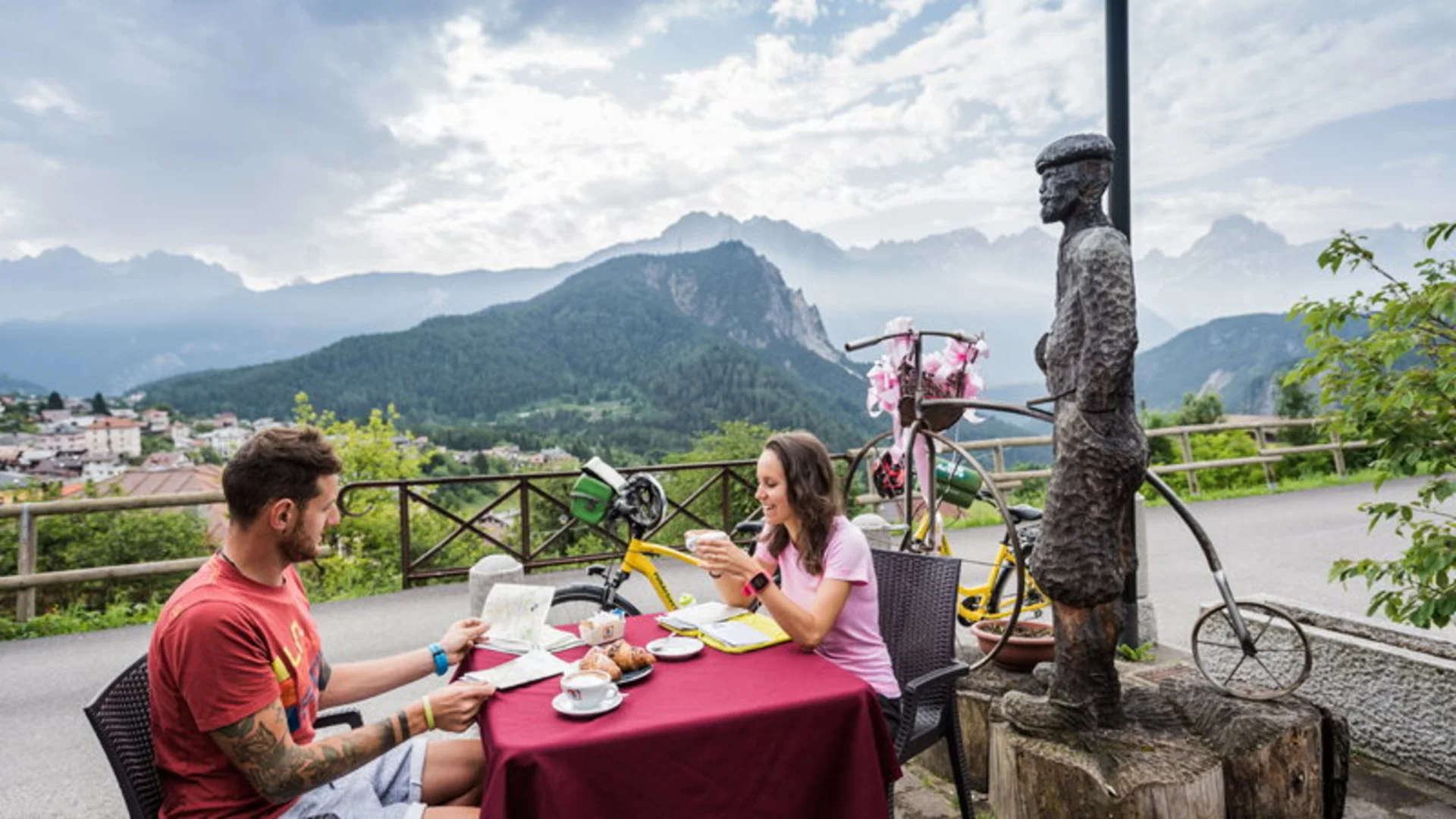 Paar trinkt Kaffee im Freien mit Bergblick und ungewöhnlicher Fahrradstatue