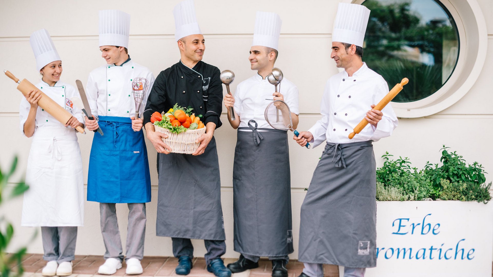 Five chefs in uniform holding cooking tools and basket of vegetables