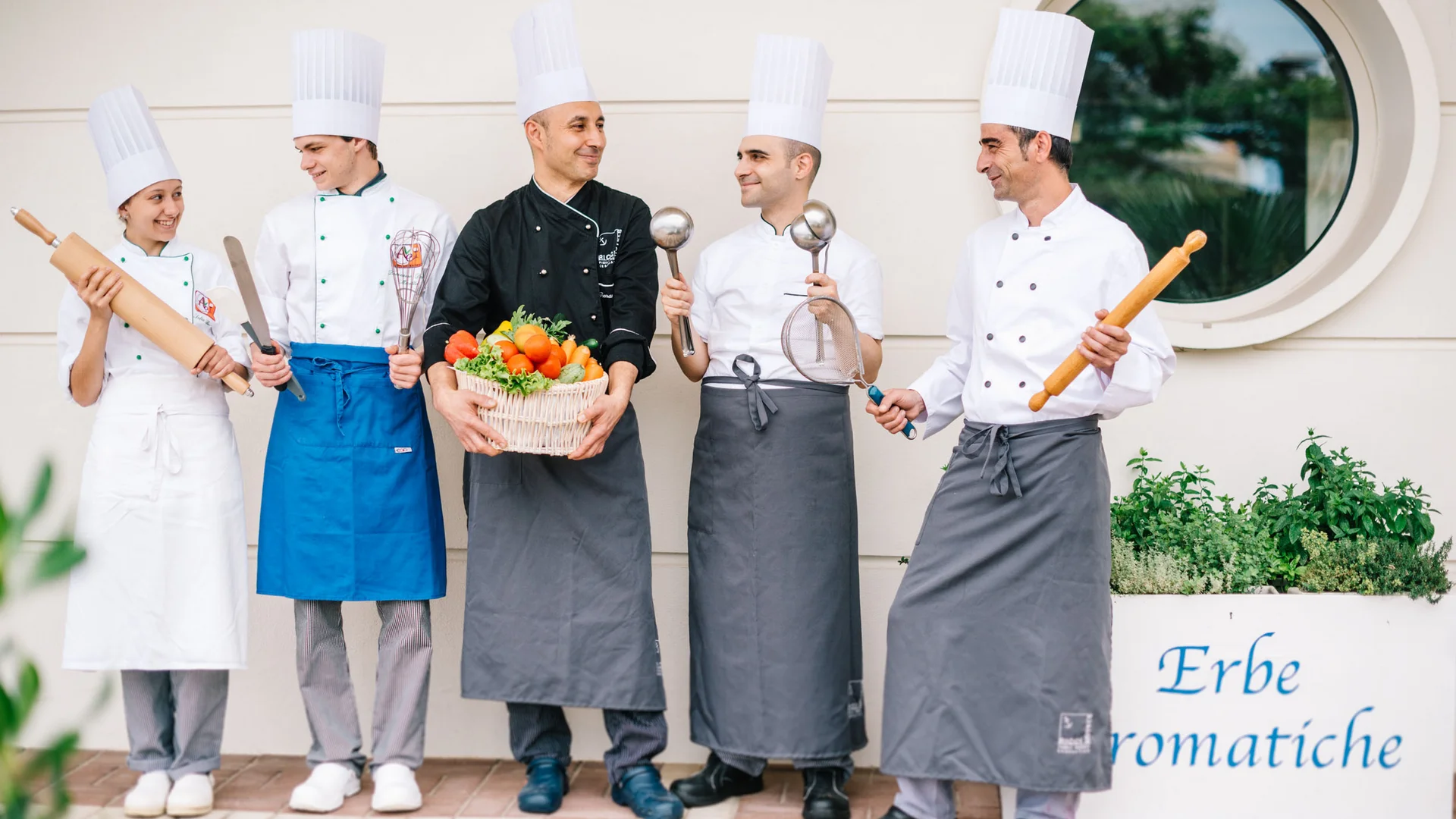 Five chefs in uniform holding cooking tools and basket of vegetables