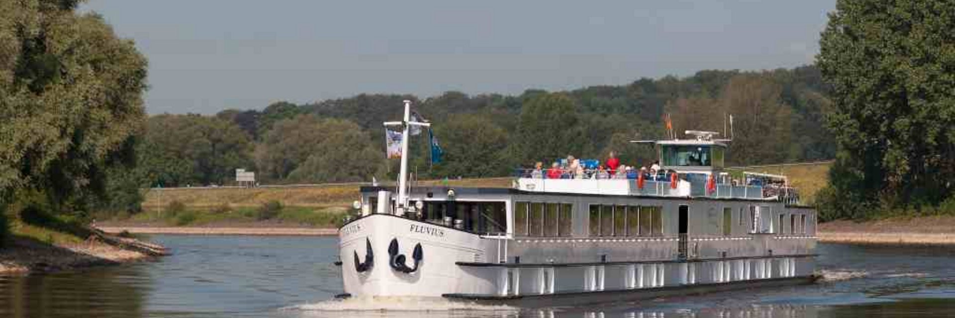 Ausflugsschiff fährt auf ruhigem Fluss mit Menschen auf Deck und Bäumen am Ufer
